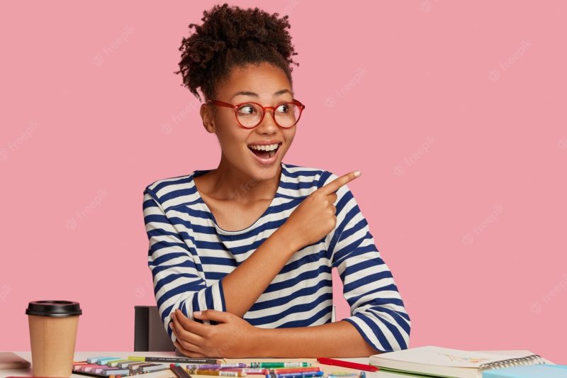 Art stationary. glad black ethnic woman with cheerful expression, wears spectacles for good vision, points at upper right corner, notices something amazing, uses notebook, colored pencils for drawing Free Photo