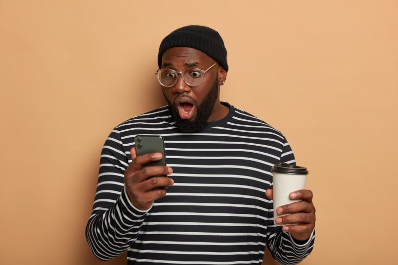 Astonished bearded young man stares at modern cellular, wears black hat and striped jumper Free Photo