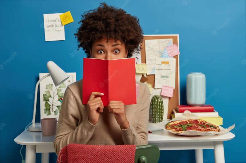 Astonished college student covers face with red diary, shocked to forget about important task to prepare Free Photo