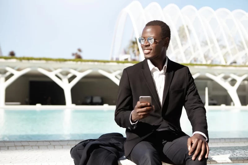 Attractive African American manager in stylish formal wear and shades using mobile phone, sitting on bench in urban setting while waiting for colleagues for lunch, smiling happily as he notices them Free Photo