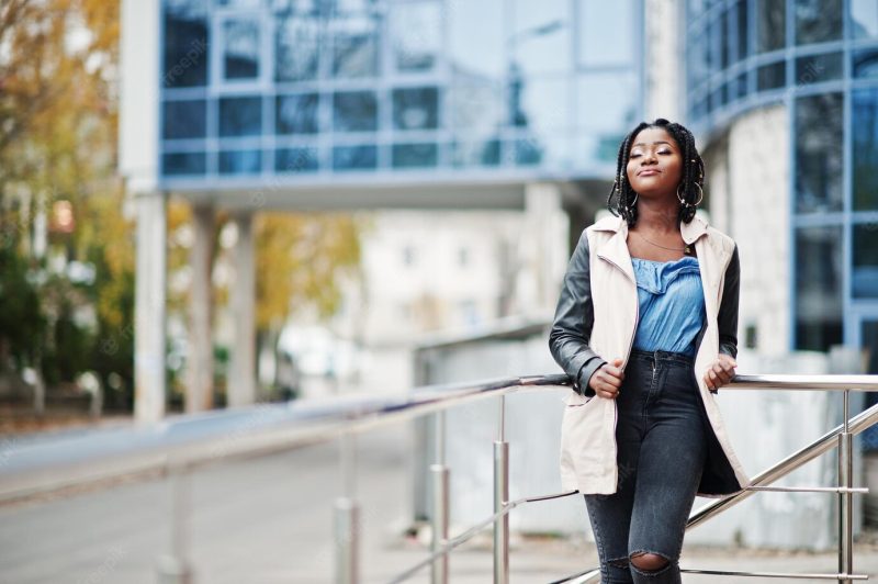 Attractive african american woman with dreads in jacket posed near railings against modern multistory building Free Photo