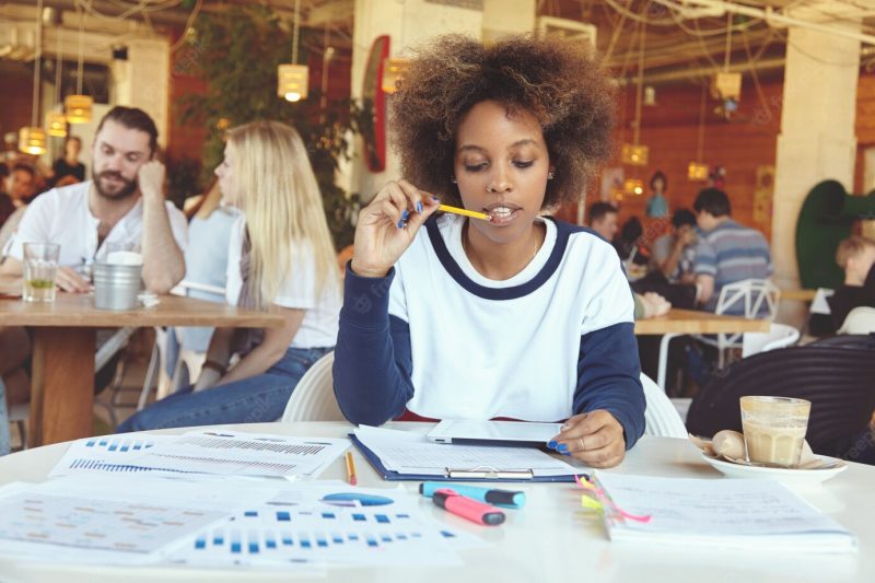 Attractive African student girl in casual clothes sitting at university canteen with touch pad pc, surfing internet while preparing for exams, touching her lips with pencil, looking thoughtful Free Photo