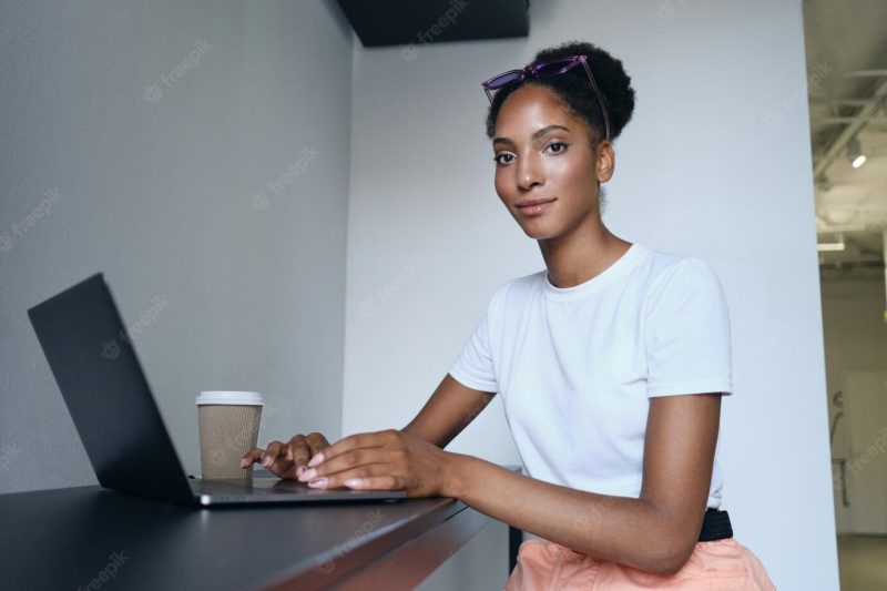 Attractive casual african american girl working on laptop with coffee to go confidently looking in camera in modern office Free Photo