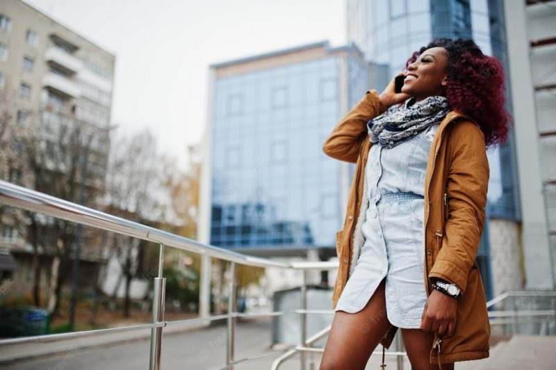 Attractive curly african american woman in brown coat posed near railings against modern multistory building speaking on mobile phone Free Photo