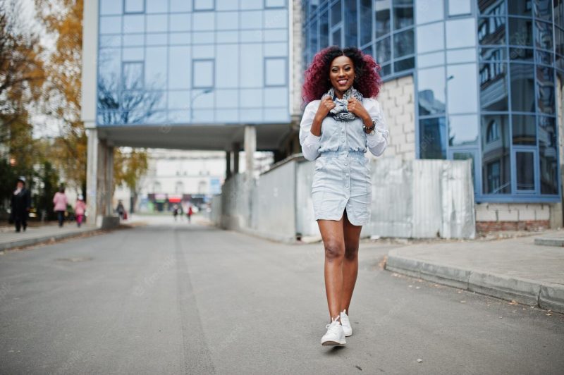 Attractive curly african american woman in jeans dress posed against modern multistory building Free Photo