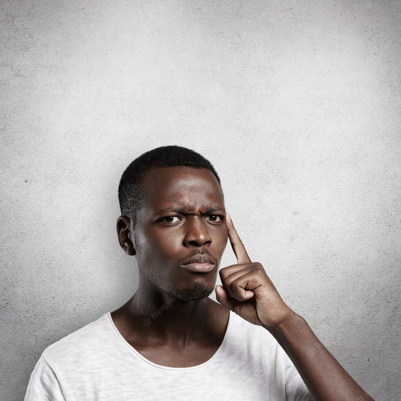 Attractive dark-skinned man wearing white t-shirt holding finger on his temple, trying hard to remember something important, frowning, looking concentrated and focused. Free Photo