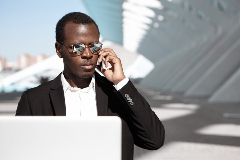 Attractive young frican American businessman wearing stylish sunglasses and formal clothing sitting at urban cafe in front of laptop, having phone conversation with partners while waiting for coffee Free Photo