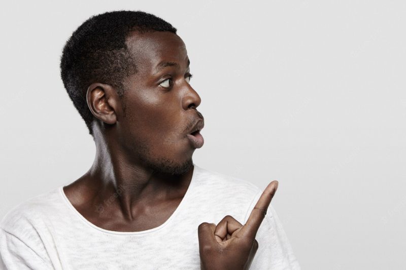 Attractive young dark-skinned customer or employee looking surprised or shocked, pointing his finger at white wall with copy space for your advertising content, saying ‘look at that!” Free Photo