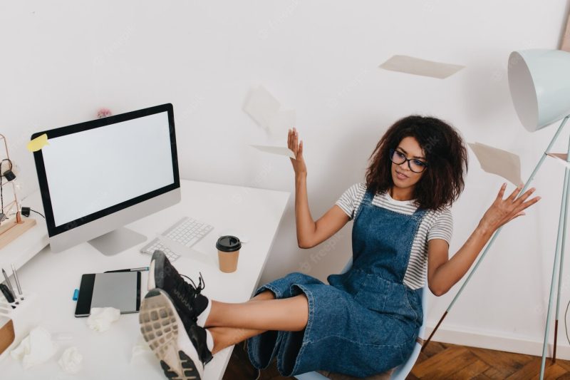 Black curly girl is scattering the documents while sitting on chair with legs on the table Free Photo