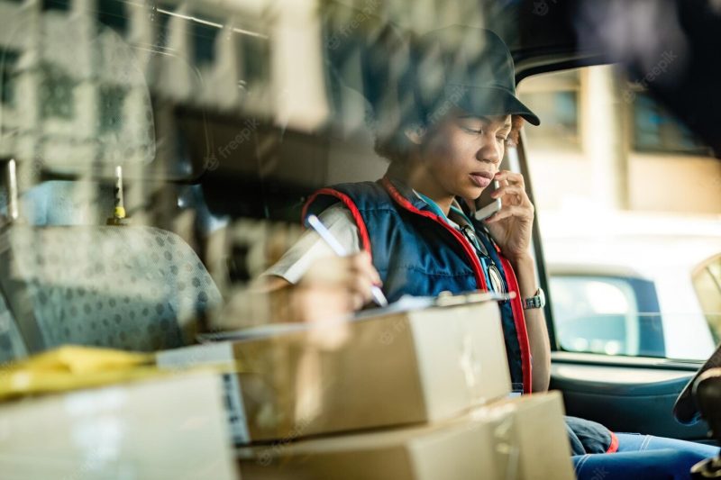 Black female courier communicating on mobile phone and taking notes while sitting in delivery van the view is through window Free Photo