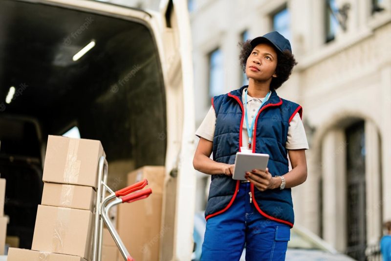 Black female courier using digital tablet while unloading packages from delivery truck on the street Free Photo