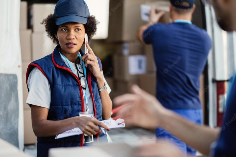 Black female deliverer communicating with coworker while talking on mobile phone and organizing package delivery Free Photo