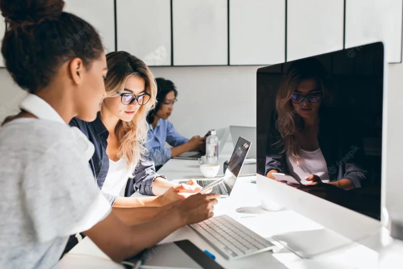 Black female it-specialist sitting beside modern computer with black screen and talking with friends. indoor portrait of busy young people working in international company. Free Photo