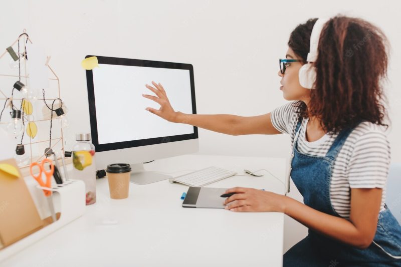 Black girl in denim sitting at the table with stationery and touching computer screen Free Photo