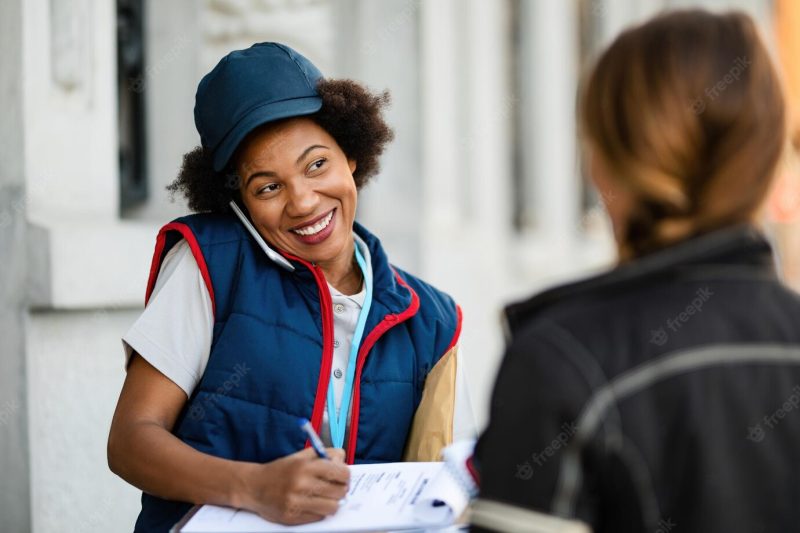 Black happy postwoman filling documents and talking on mobile phone while delivering package to a customer Free Photo