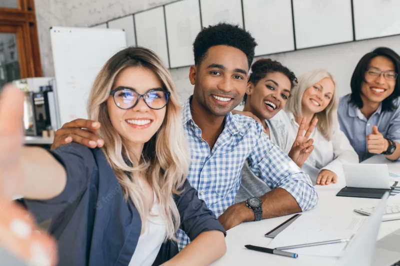 Black office worker in checkered shirt embracing blonde secretary woman while she making selfie. young managers of international company having fun during meeting. Free Photo