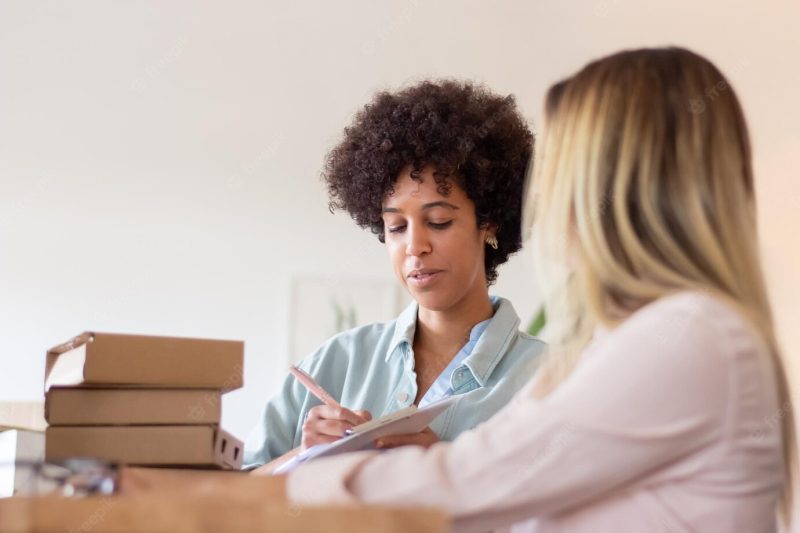 Blackwoman discussing parcel with colleague and making note in clipboard. warehouse workers preparing goods for dispatch. logistics concept Free Photo