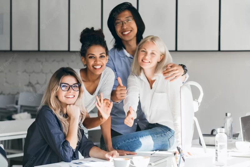 Blissful asian programmer embracing female blonde colleague for photo and smiling. indoor portrait of glad african office worker posing beside European friend at her workplace. Free Photo