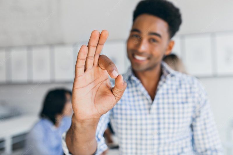 Blissful office worker having fun with colleagues and showing okay sign. indoor portrait of laughing black young man working in international company with his hand in focus. Free Photo