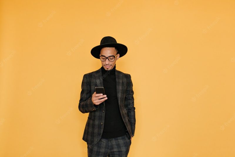 Bored African man in fashionable costume standing on yellow wall with phone. indoor portrait of joyful black boy wears gray clothes. Free Photo