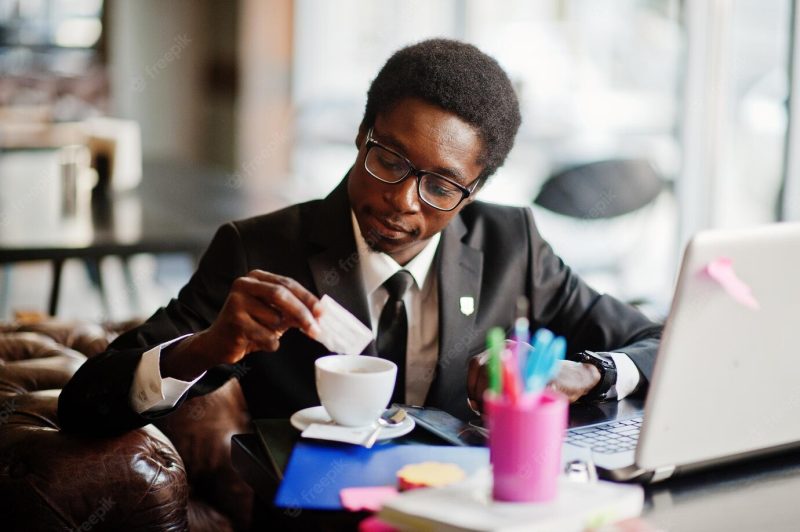 Business african american man wear on black suit and glasses sitting at office with laptop and working pours sugar into coffee Free Photo