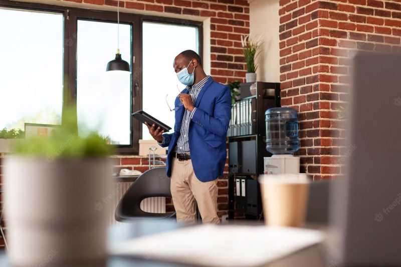 Business man wearing face mask and using digital tablet to plan company project. employee standing in startup office and working on device with technology during coronavirus pandemic. Free Photo