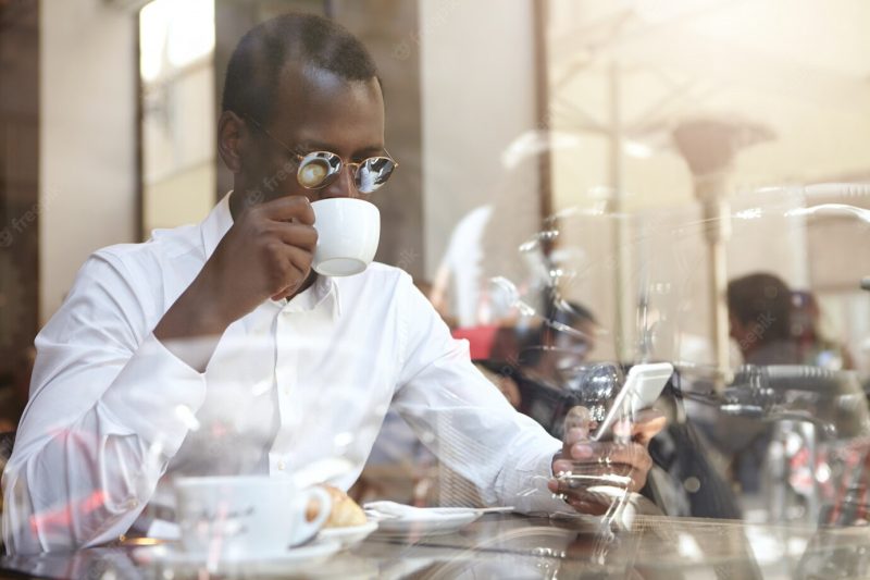 Business, modern technology, communication and people concept. confident stylish african american businessman in round sunglasses drinking cappccino at coffee shop, sitting by window with cell phone Free Photo