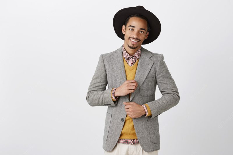 Businessman on party, celebrating successful deal. confident charming african-american entrepreneur in stylish formal clothes and hat, checking outfit and smiling broadly, being sassy over gray wall Free Photo