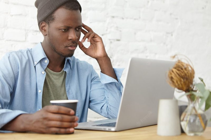 Picture of fashionable young African American male programmer, blogger, freelancer or startupper using laptop computer at coworking space for remote work and drinking coffee, having happy look Free Photo