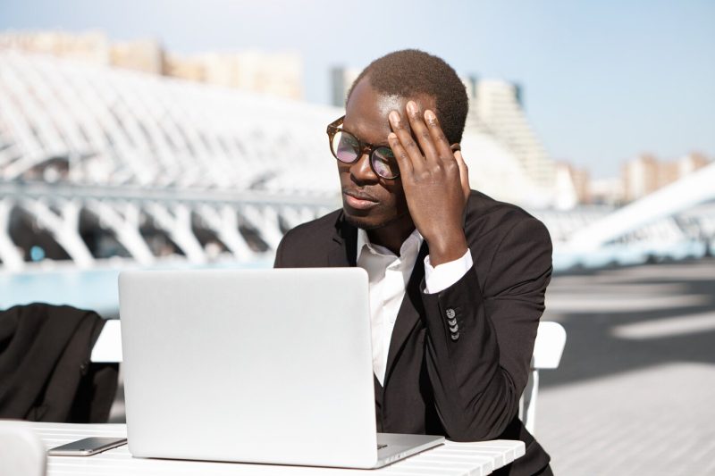 Candid shot of unhappy young African American manager feeling tired and frustrated, sitting at urban cafe with generic laptop pc, touching head, trying to concentrate on work, looking exhausted Free Photo