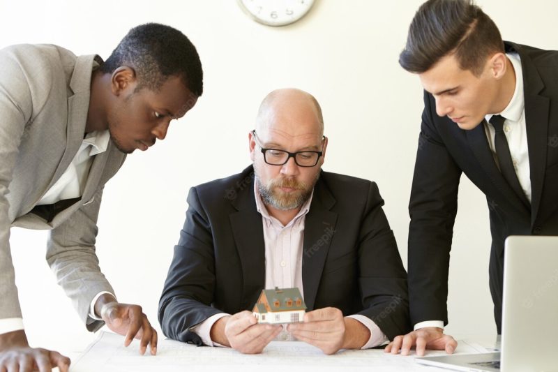 Caucasian boss in glasses holding scale model house of future real estate while two young architects presenting construction project to him. Free Photo