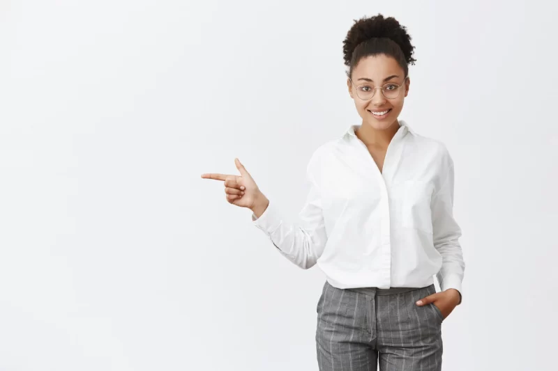 Charming and friendly office worker showing way out to customer. portrait of polite smart and creative female employer in glasses and pants, holding hand in pocket, pointing left, indicating at exit Free Photo