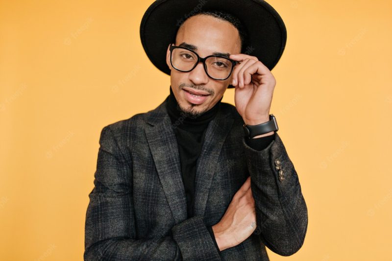 Charming young man with brown skin posing in elegant wristwatch. serious african guy in trendy suit standing on yellow wall. Free Photo