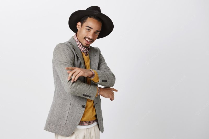 Check out my new outfit. portrait of stylish confident dark-skinned male student in trendy hat, making cool gestures, dancing or posing to show clothes, smiling Free Photo