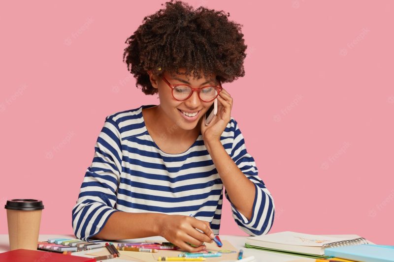 Cheerful black lady enjoys painting, makes picture on blank sheet of paper, wears optical glasses, has telephone conversation, smiles gently as discusses something pleasant, isolated on pink wall Free Photo