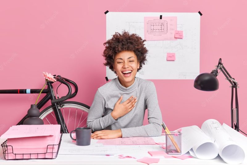 Cheerful dark skinned inspired female design worker giggles happily sits at workplace makes drawings expresses positive emotions enjoys her occupation studies technical blueprints at desktop Free Photo