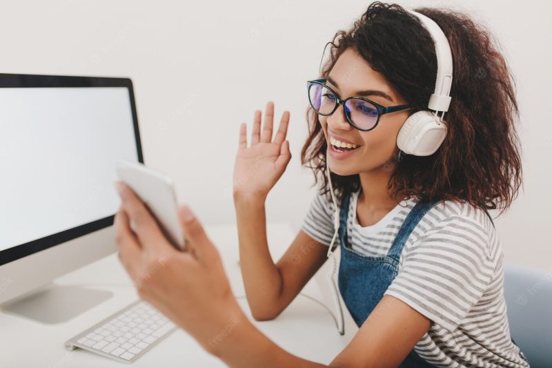 Cheerful girl in stylish shirt and headphones communicates with friend by video link Free Photo