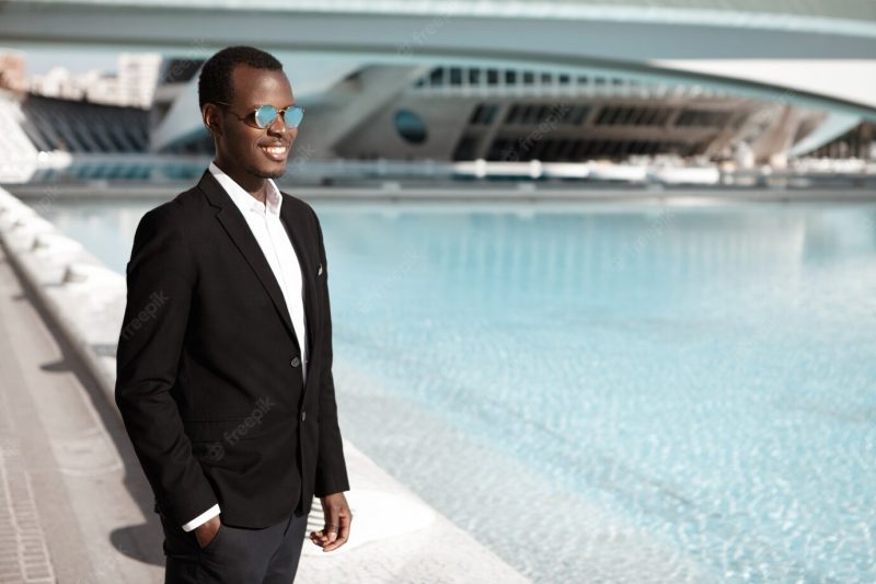 Cheerful happy young African American office worker wearing black elegant suit and stylish shades standing at urban fountain keeping hand inside pocket Free Photo