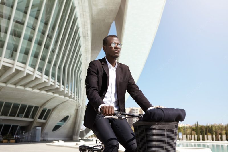 Cheerful stylish black European male businessman using bicycle to get to office while his car is broken, cycling in urban setting, passing by modern buildings and fountain on sunny summer day Free Photo