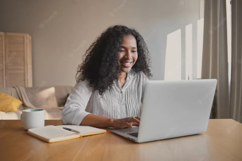 Cheerful young african american woman copywriter sitting in front of open laptop with mug and copybook on desk, feeling inspired, working on new motivation article. people, occupation and creativity Free Photo