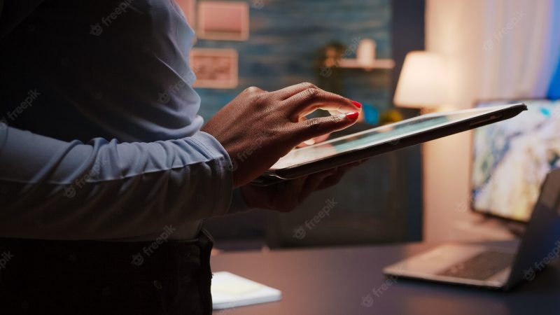 Close-up photo of black female hands holding tablet computer standing in living room late at night. african american woman using social network, texting and blogging working overtime for job Free Photo