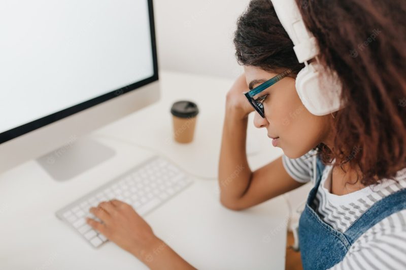 Close-up portrait of tired black girl in big white headphones sitting at workplace and typing on keyboard Free Photo