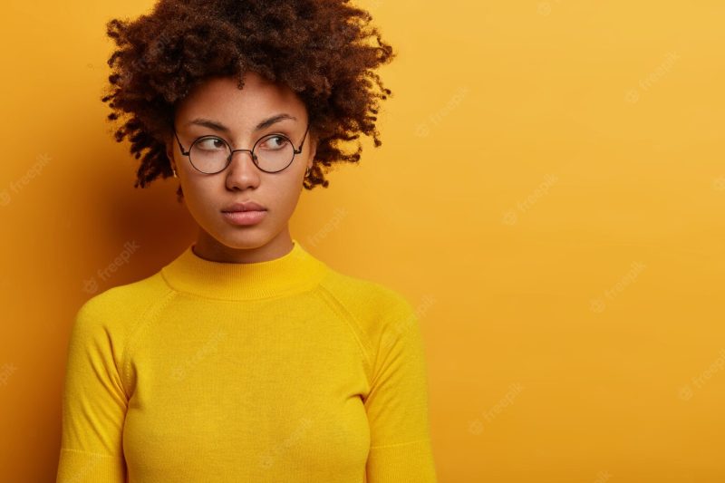 Close up shot of contemplative young female model wears round spectacles ad yellow clothes, looks aside with pensive expression, thinks over plan, poses indoor, blank space for your advertisement Free Photo