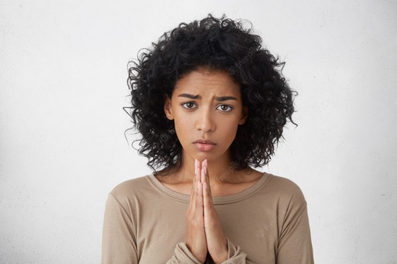 Close up shot of cute young dark-skinned lady wearing casual long sleeved top pressing palms together, feeling worried and desperate while praying for wellness and good health of her sick husband Free Photo