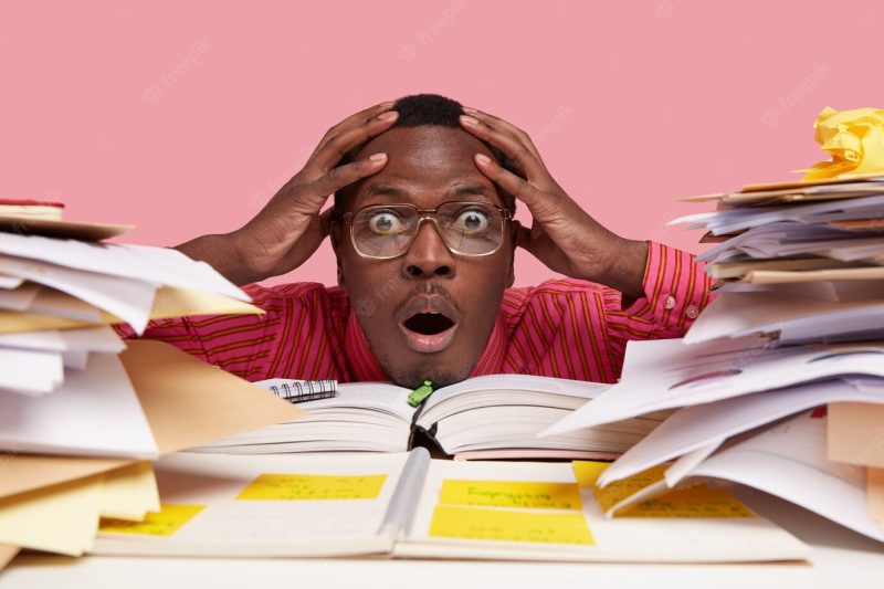 Close up shot of shocked stupefied black african american man keeps both hands on head, looks through pile of books and papers, has difficult task Free Photo