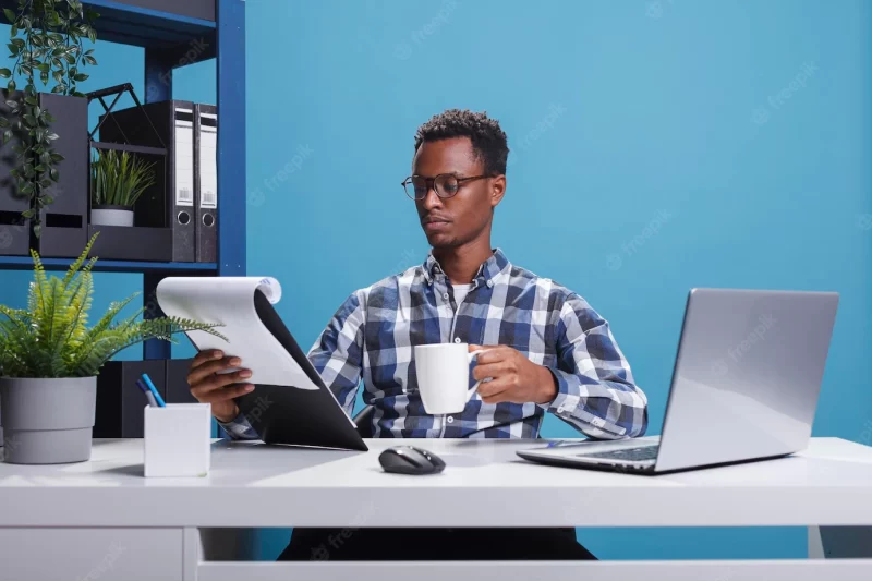 Company office worker having documentation clipboard and coffee cup looking over project management charts. young african american employee sitting in modern workspace while reviewing paperwork. Free Photo