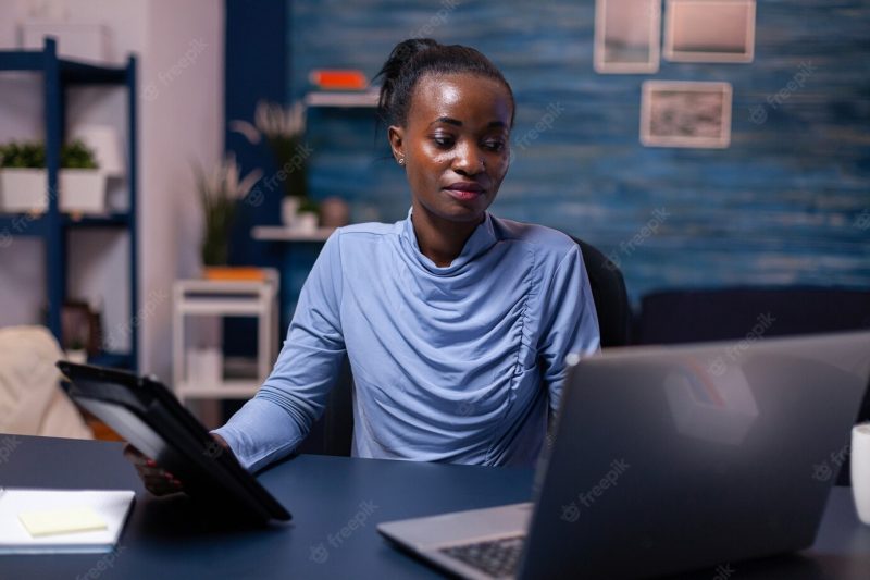 Concentrated african woman working on deadline using tablet pc and laptop in home office late at night. busy focused employee using modern technology network wireless doing overtime writing. Free Photo