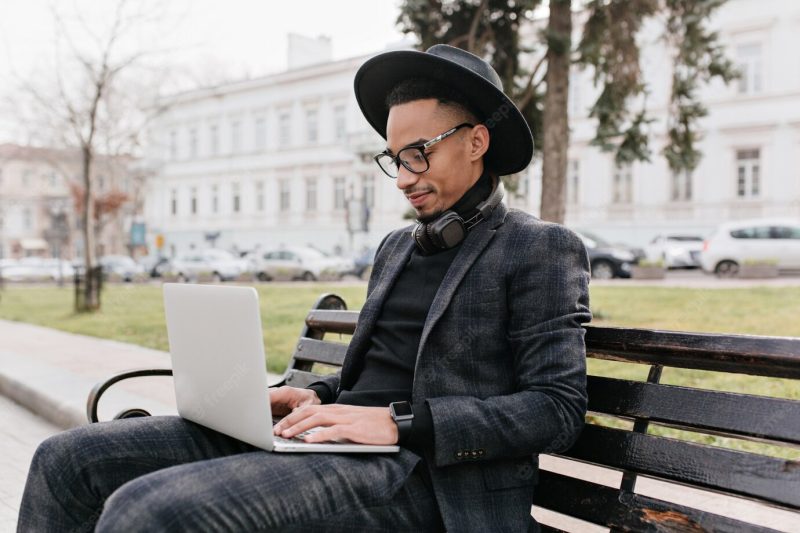 Concentrated freelance worker in hat sitting in park with computer. outdoor photo of handsome african young man typing on keyboard on nature. Free Photo