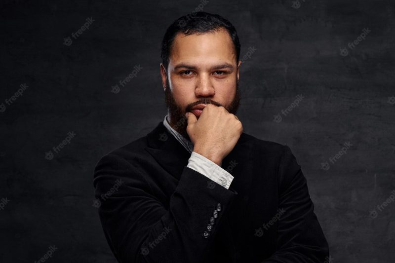 Confident african-american businessman in a black classic jacket, thinking about business while holding hand on chin. isolated on a dark background. Free Photo