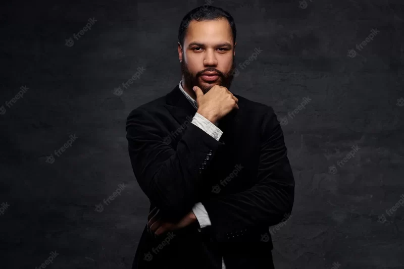 Confident african-american businessman in a black classic jacket, thinking about business while holding hand on chin. isolated on a dark background. Free Photo